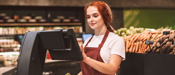 Young smiling seller in apron behind counter with vegetables weighing bananas on scale in modern supermarket